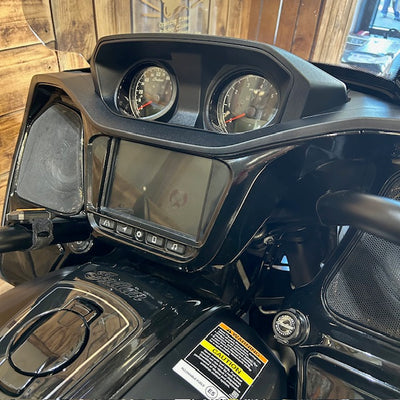 Close-up of a black motorcycle or side-by-side vehicle dashboard with two analog gauges, a central display screen, handlebars, and a wood-paneled interior background.