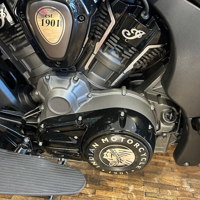 Close-up of a motorcycle engine and side cover, showing black and silver components with a circular Indian Motorcycle emblem on the side cover and a gold-toned 'est. 1901' badge.