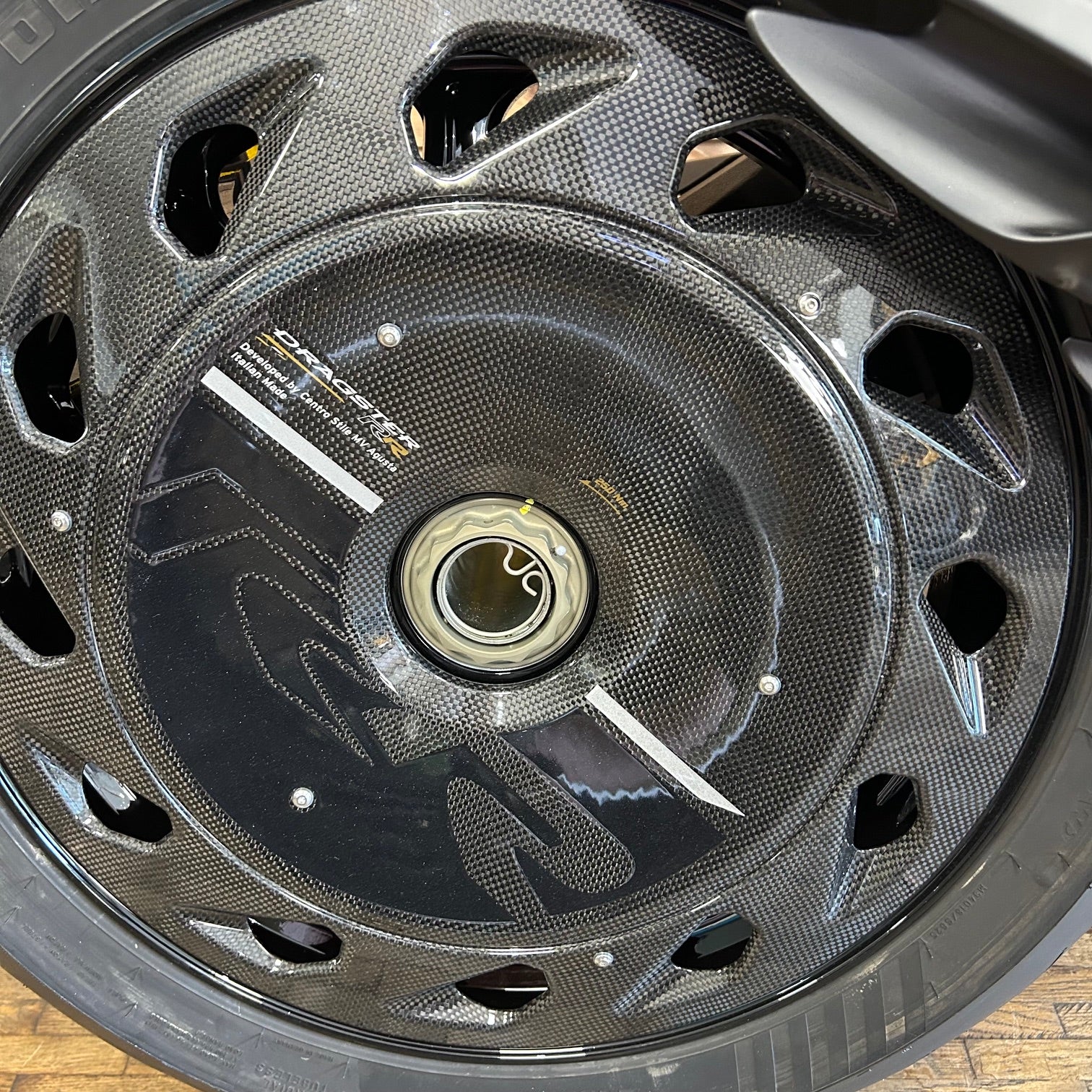 Close-up of a carbon fiber motorcycle wheel with a glossy black finish, featuring cutouts and a central hub.