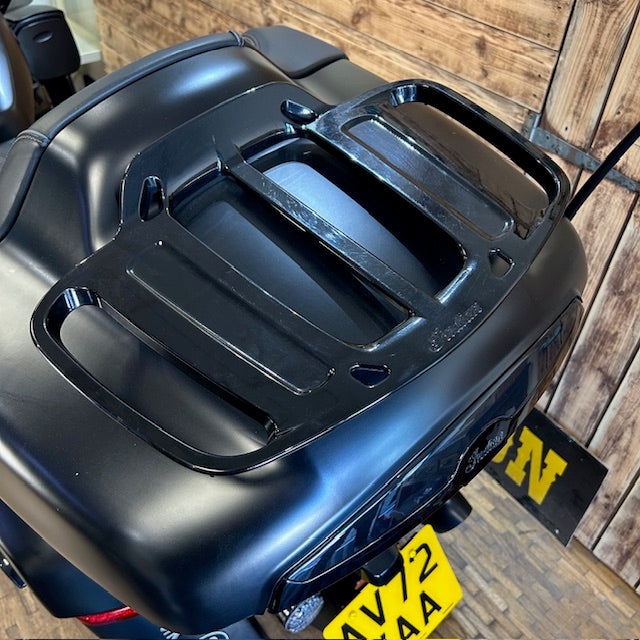 A glossy black motorcycle luggage rack mounted on the rear of a black motorcycle, photographed in a real indoor setting with wood paneling and a yellow license plate visible.