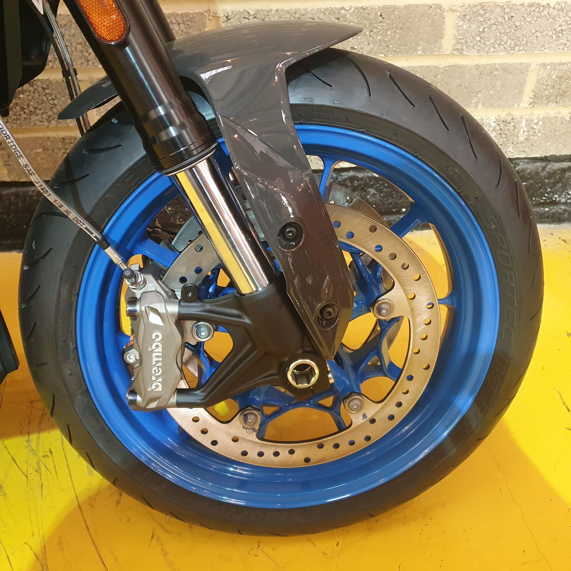 Close-up of a motorcycle wheel with blue rim and yellow suspension on a yellow surface.