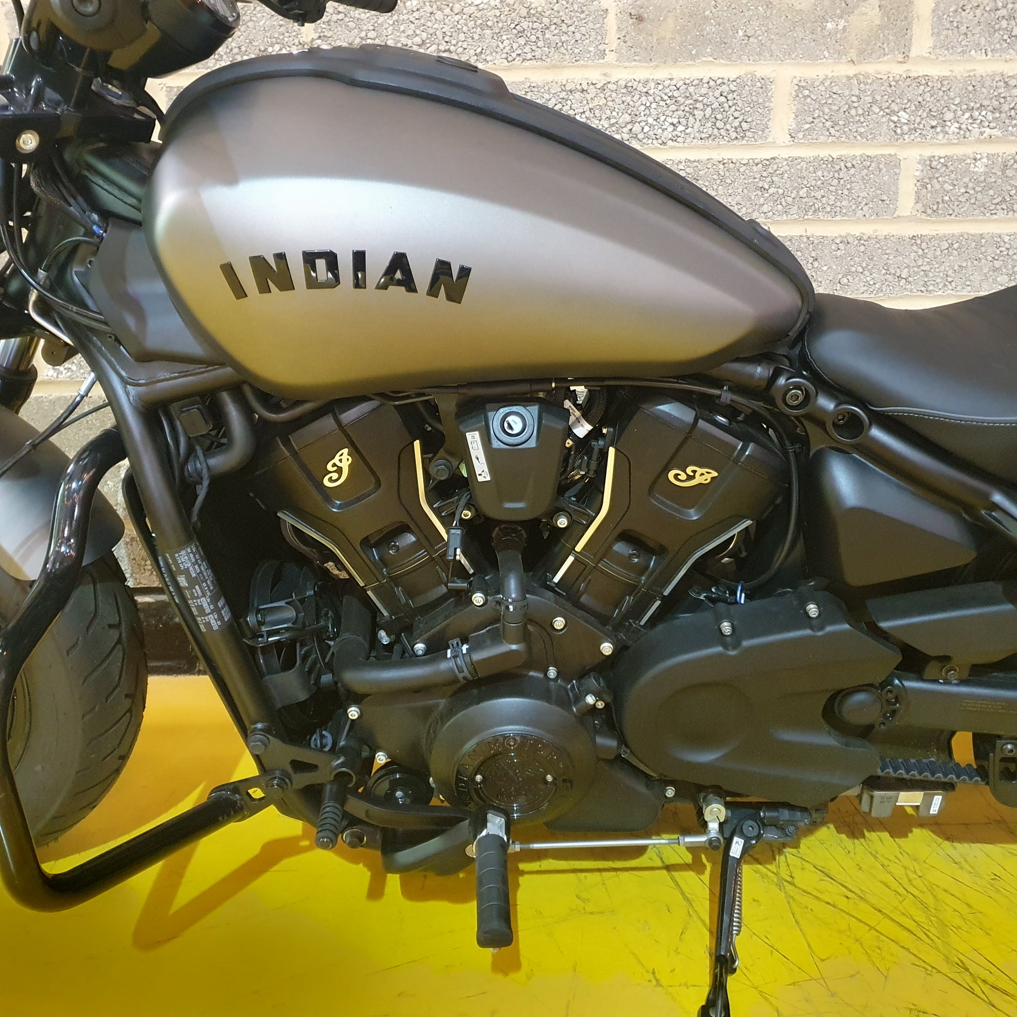 Close-up of an Indian motorcycle engine and fuel tank on a yellow surface with a brick wall background.