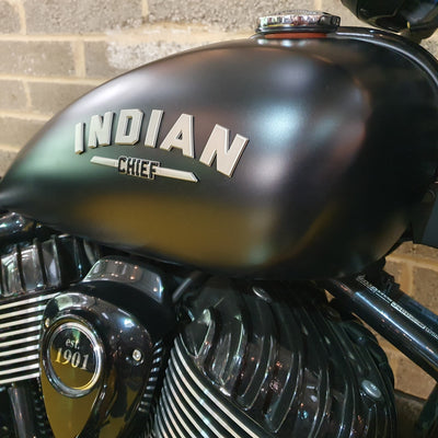 Close-up of a motorcycle fuel tank with 'Indian Chief' branding against a brick wall.