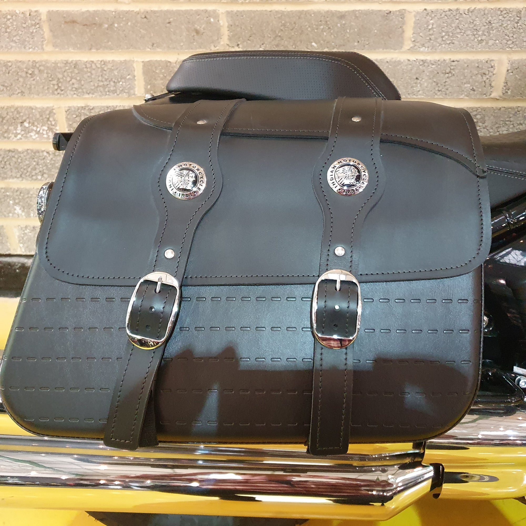 Black leather saddlebags with silver buckles on a motorcycle against a brick wall.