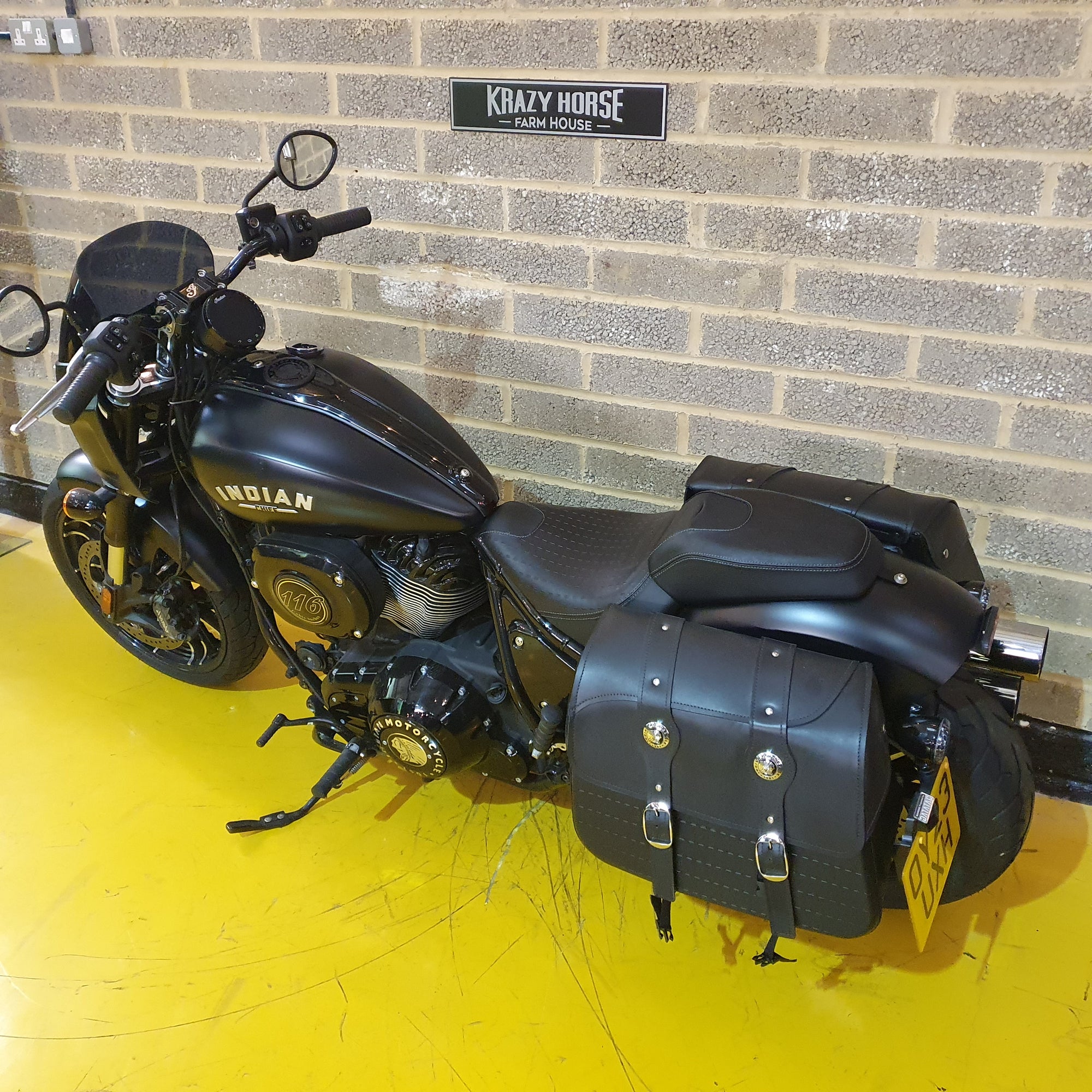Black Indian motorcycle with saddlebags on a yellow floor against a brick wall.