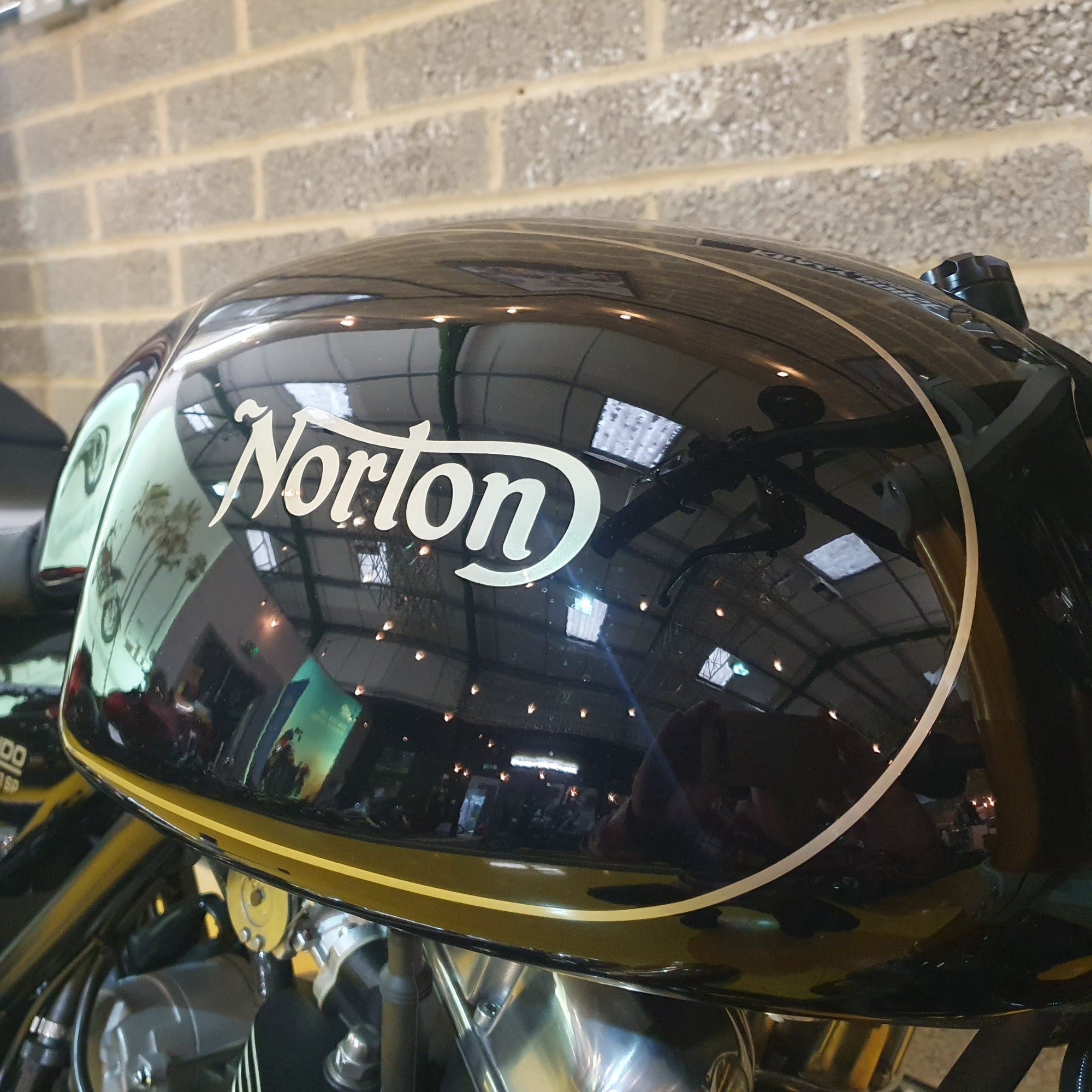Norton motorcycle fuel tank with 'Norton' branding against a brick wall.