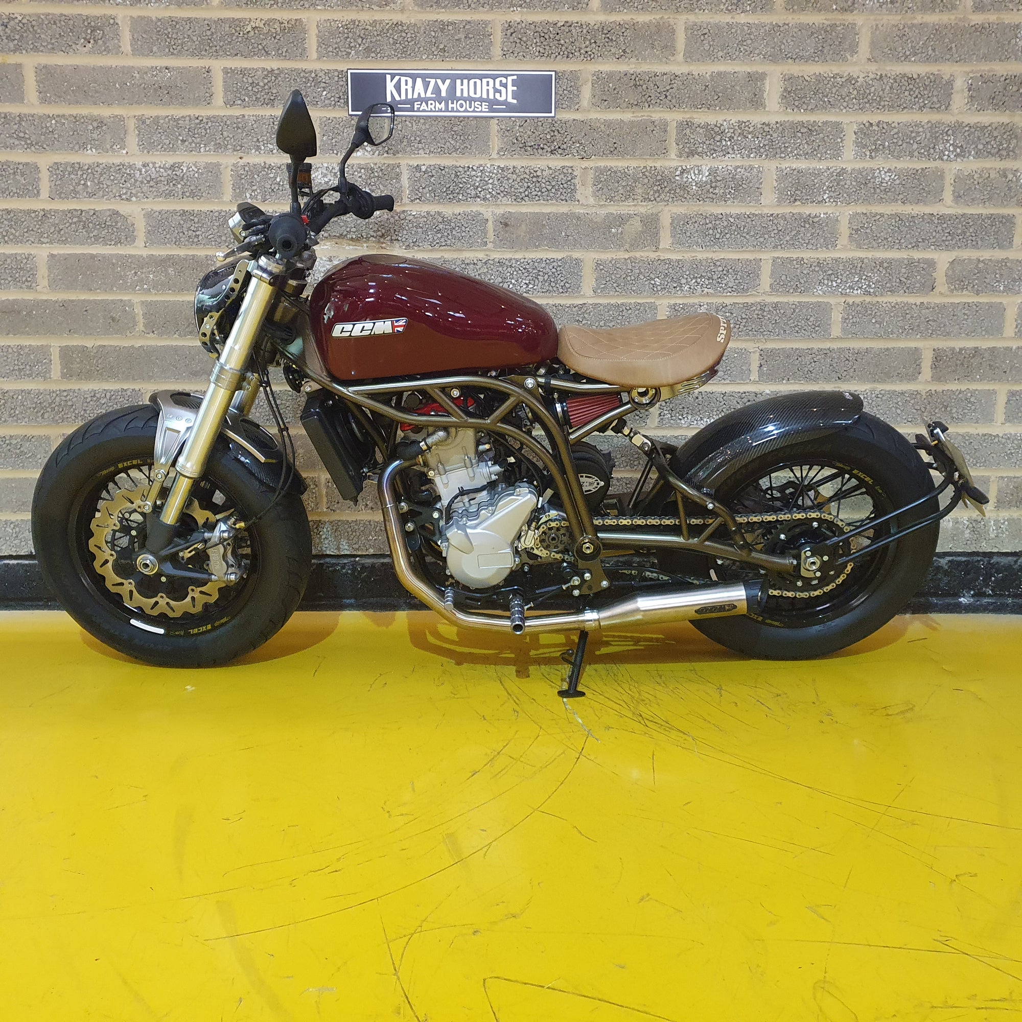 Motorcycle with maroon seat and gold accents on a yellow floor against a brick wall.