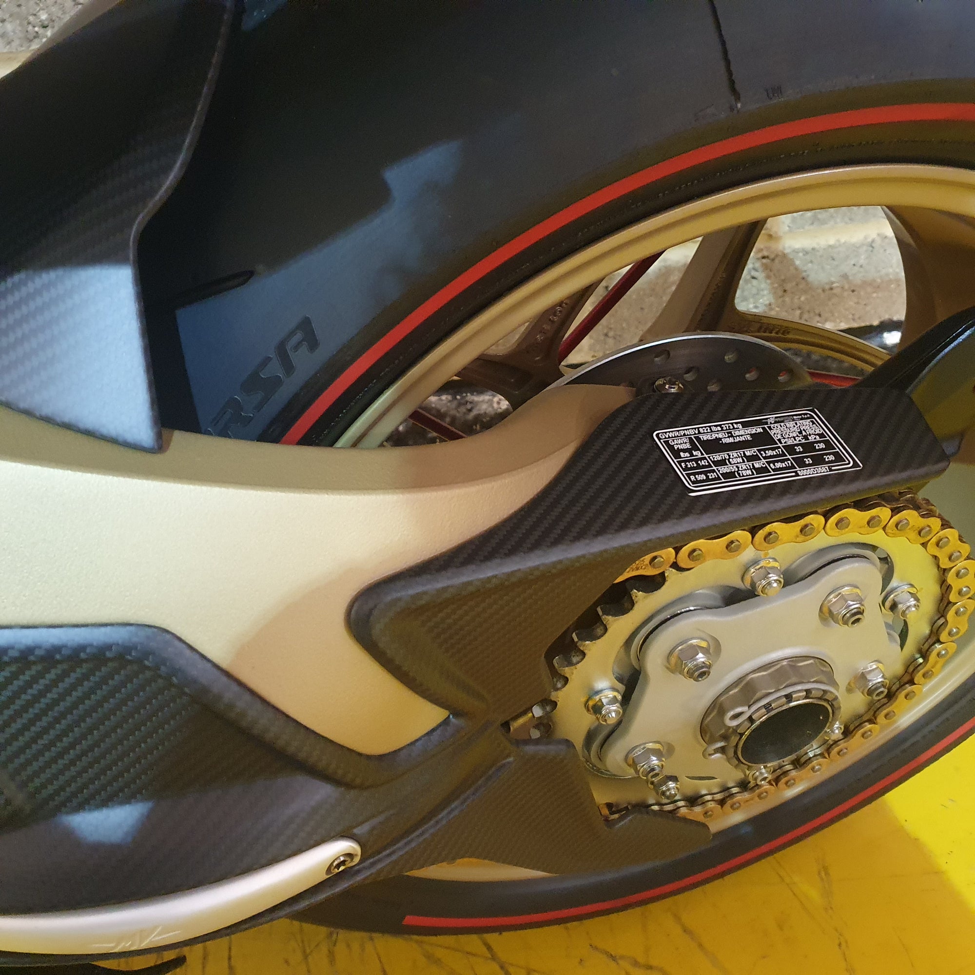 Close-up of a motorcycle chain and sprocket with a brand logo in the background.