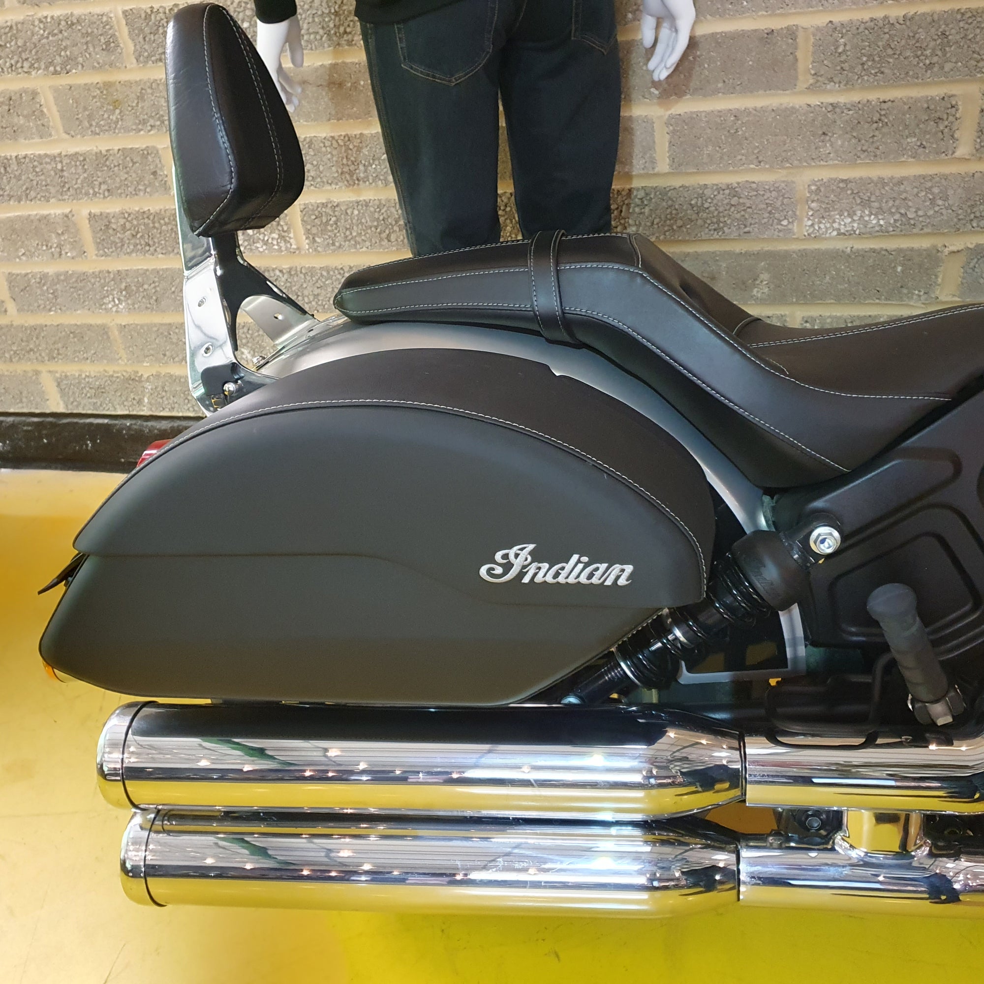 Motorcycle seat with 'Indian' logo on a yellow surface against a brick wall.