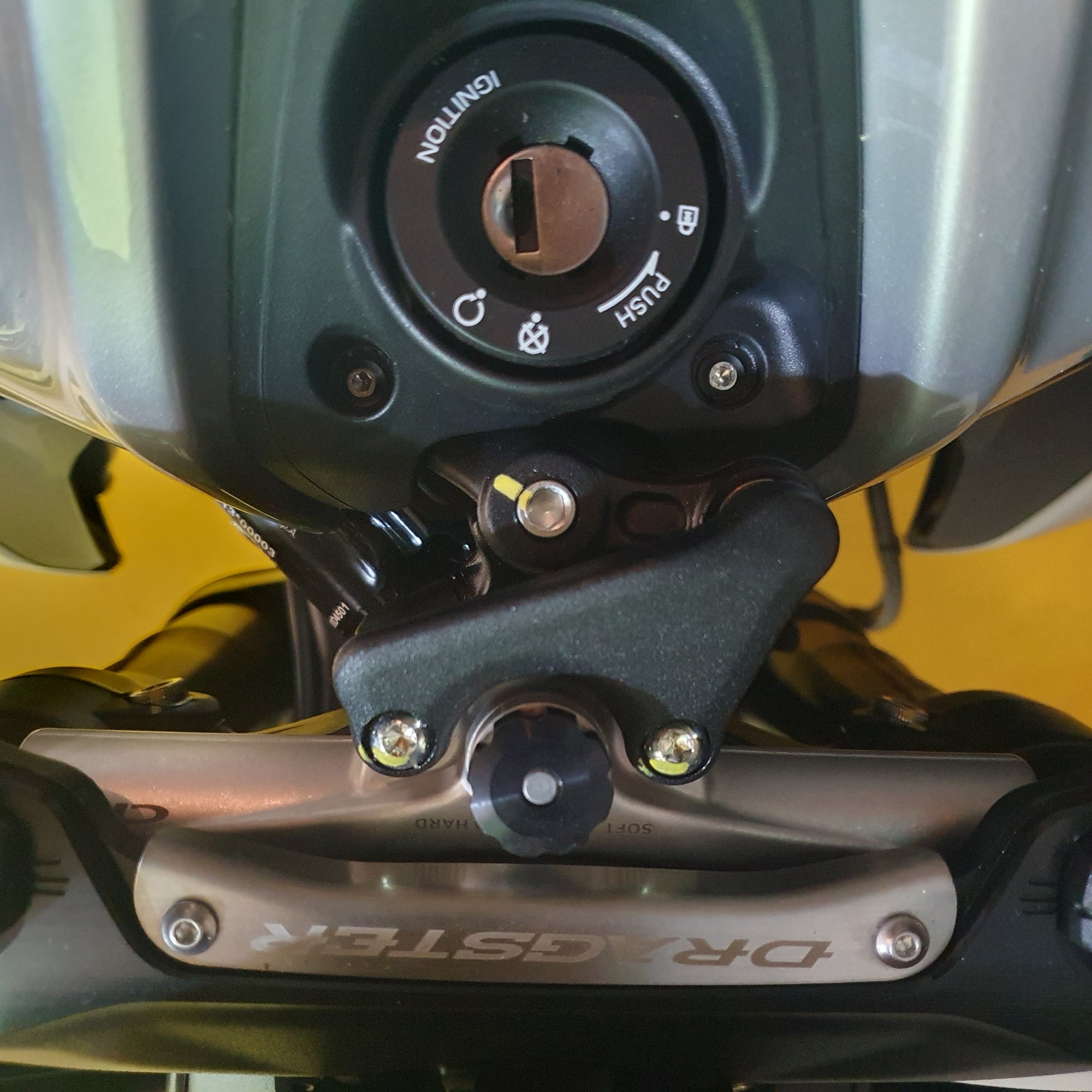 Close-up of a motorcycle fuel tank ignition switch and mounting hardware, with a silver DRAGSTER-branded metal bar visible below.
