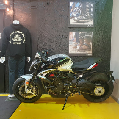 A black and silver MV Agusta motorcycle displayed indoors on a yellow floor mat against a dark textured wall, with a mannequin wearing a black hoodie on the left and framed boots photos on the wall.