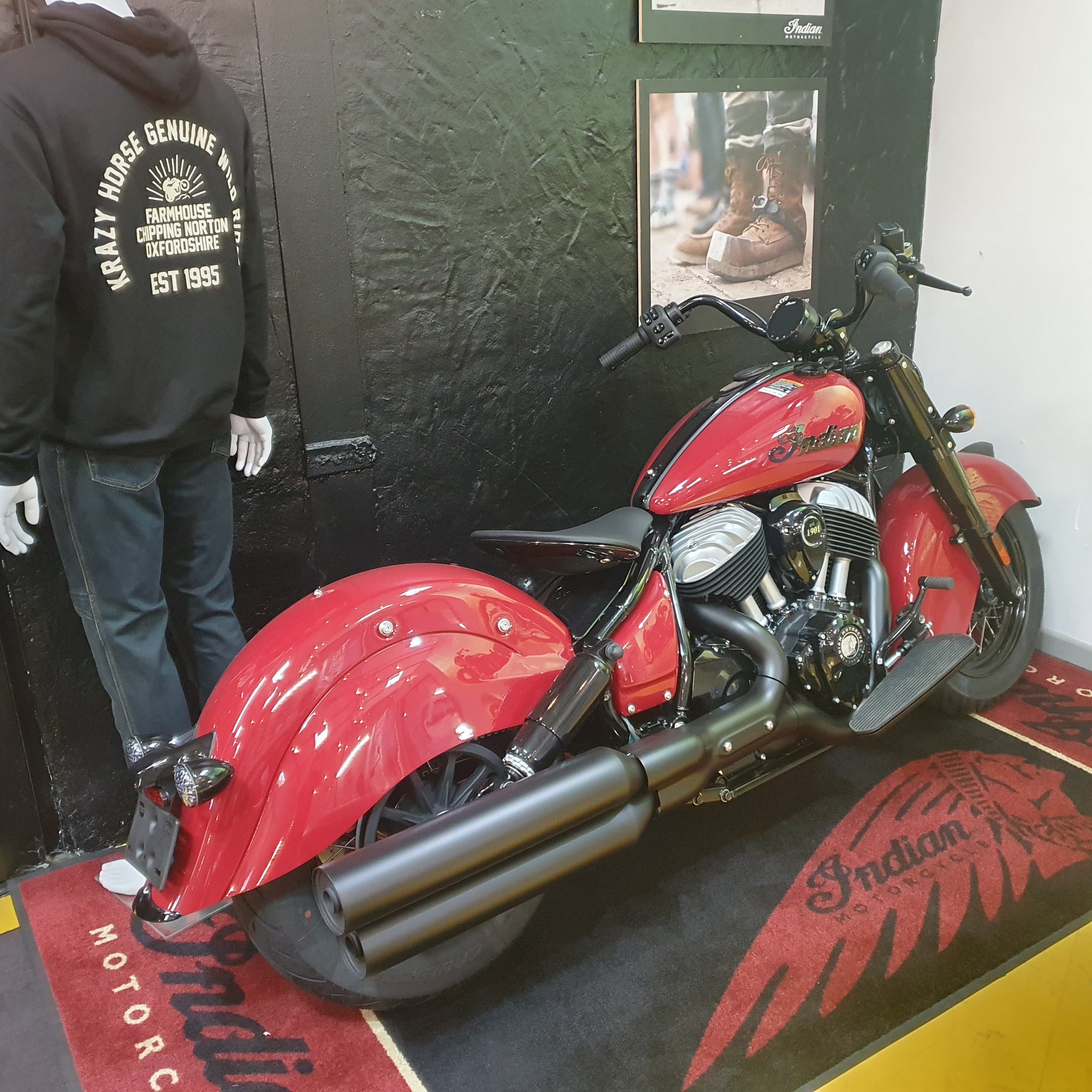 A red Indian chopper motorcycle displayed indoors against a dark wall, with a mannequin wearing a black hoodie standing beside it and a branded floor mat beneath the bike.