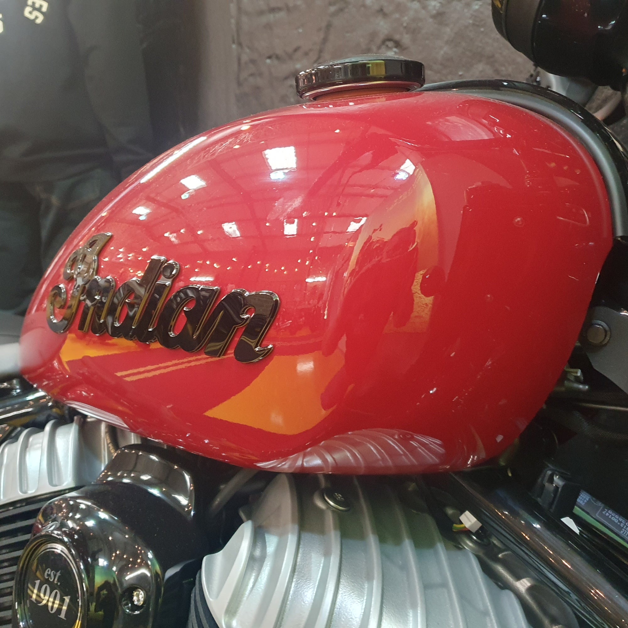 Close-up of a red Indian motorcycle fuel tank with chrome engine parts and the Indian logo on a workshop or garage background.