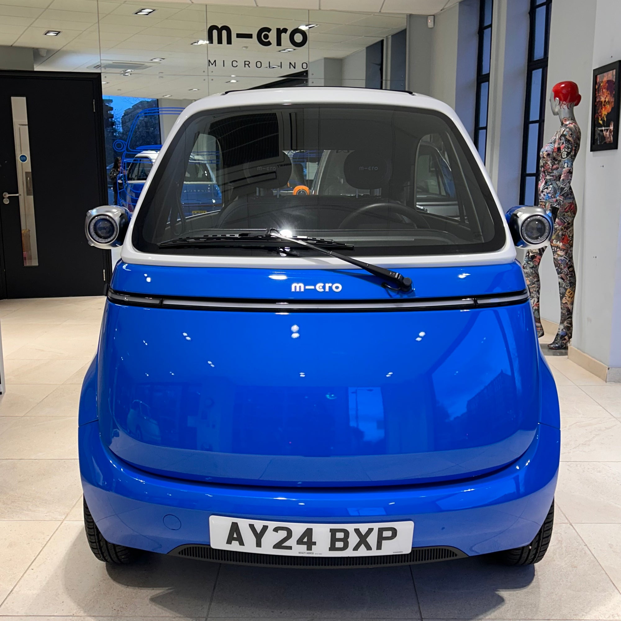 Blue microcar with 'm-cro' branding inside a showroom.