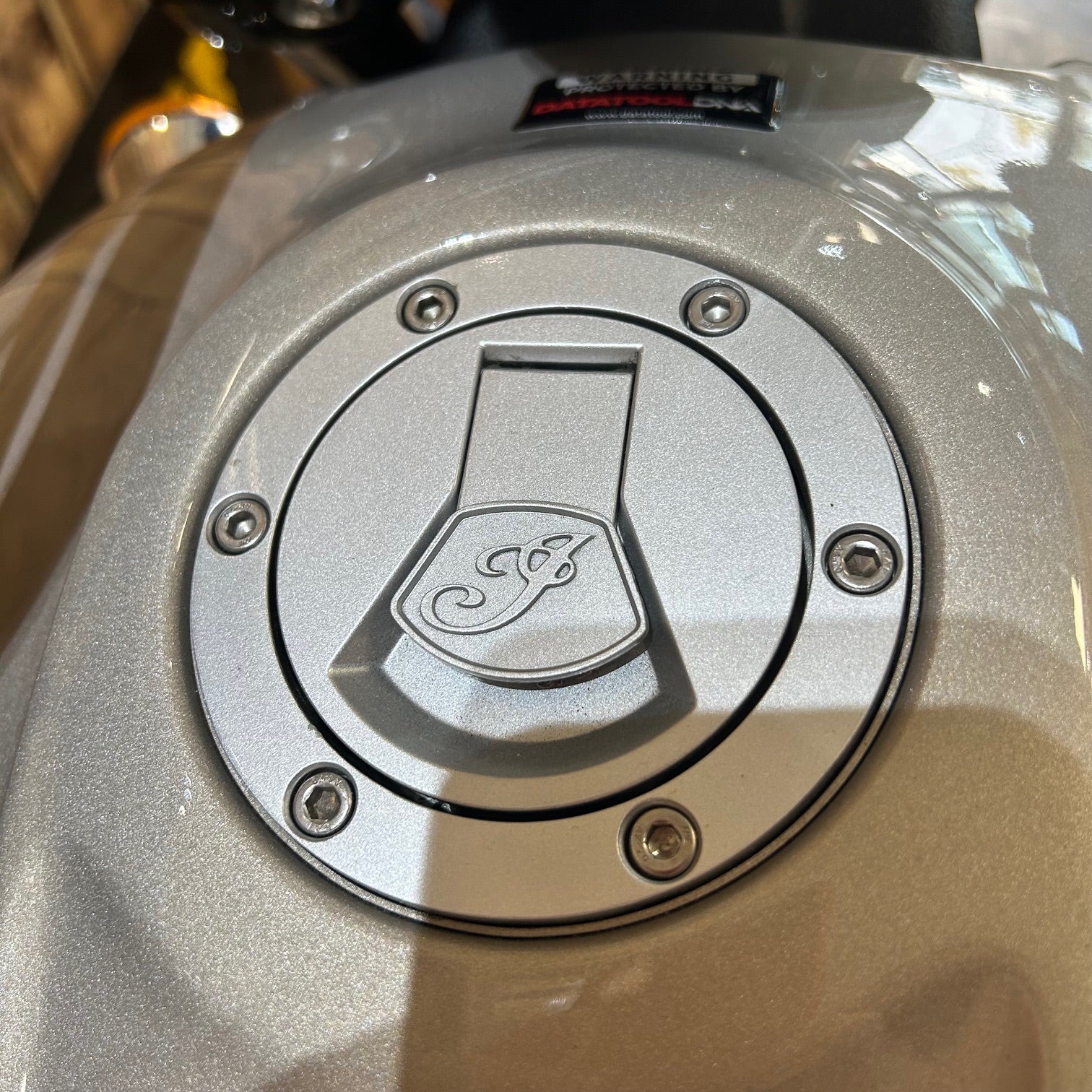 Close-up of a metallic fuel cap with a logo on a motorcycle.