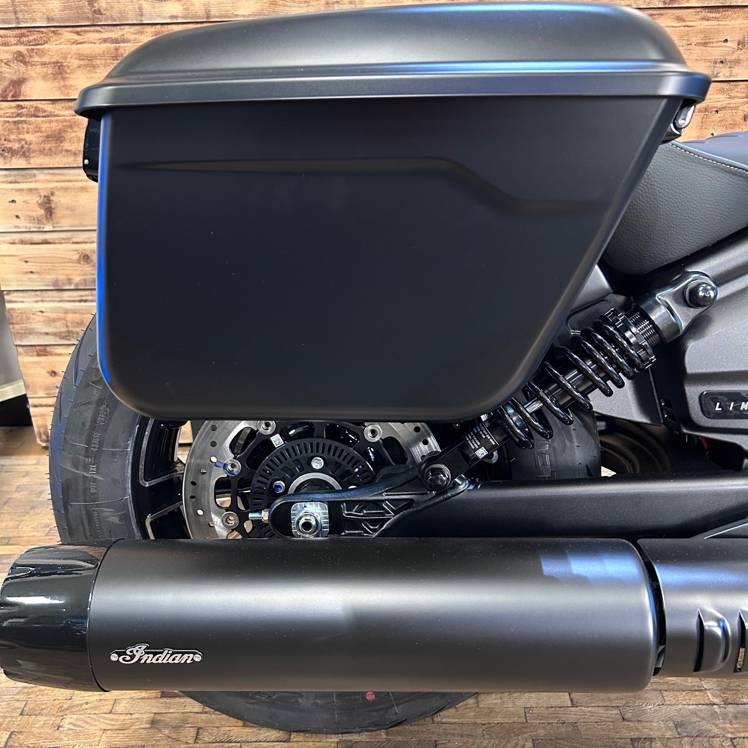 Close-up of a motorcycle with a black exhaust and saddlebag on a wooden floor.