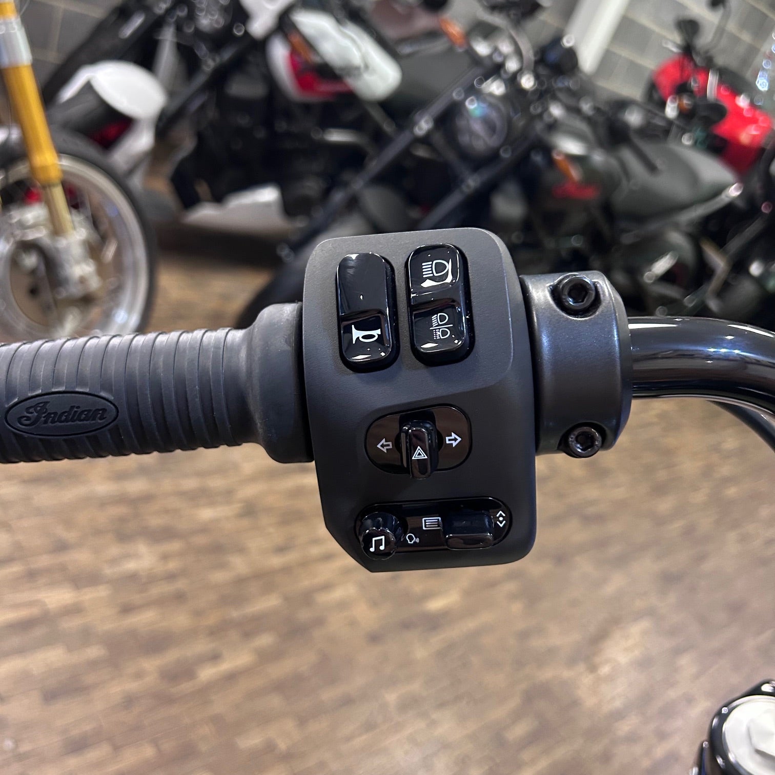Close-up of motorcycle handlebar controls with 'Indian' branding, blurred background of motorcycles.