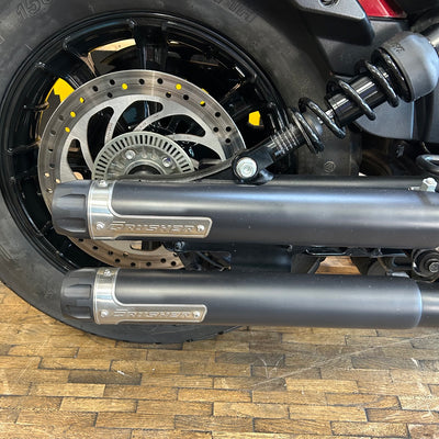 Close-up of a motorcycle wheel with 'Crusher' exhaust on a wooden floor.