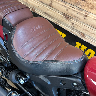 Brown leather seat of a motorcycle with 'Indian' branding against a wooden background.