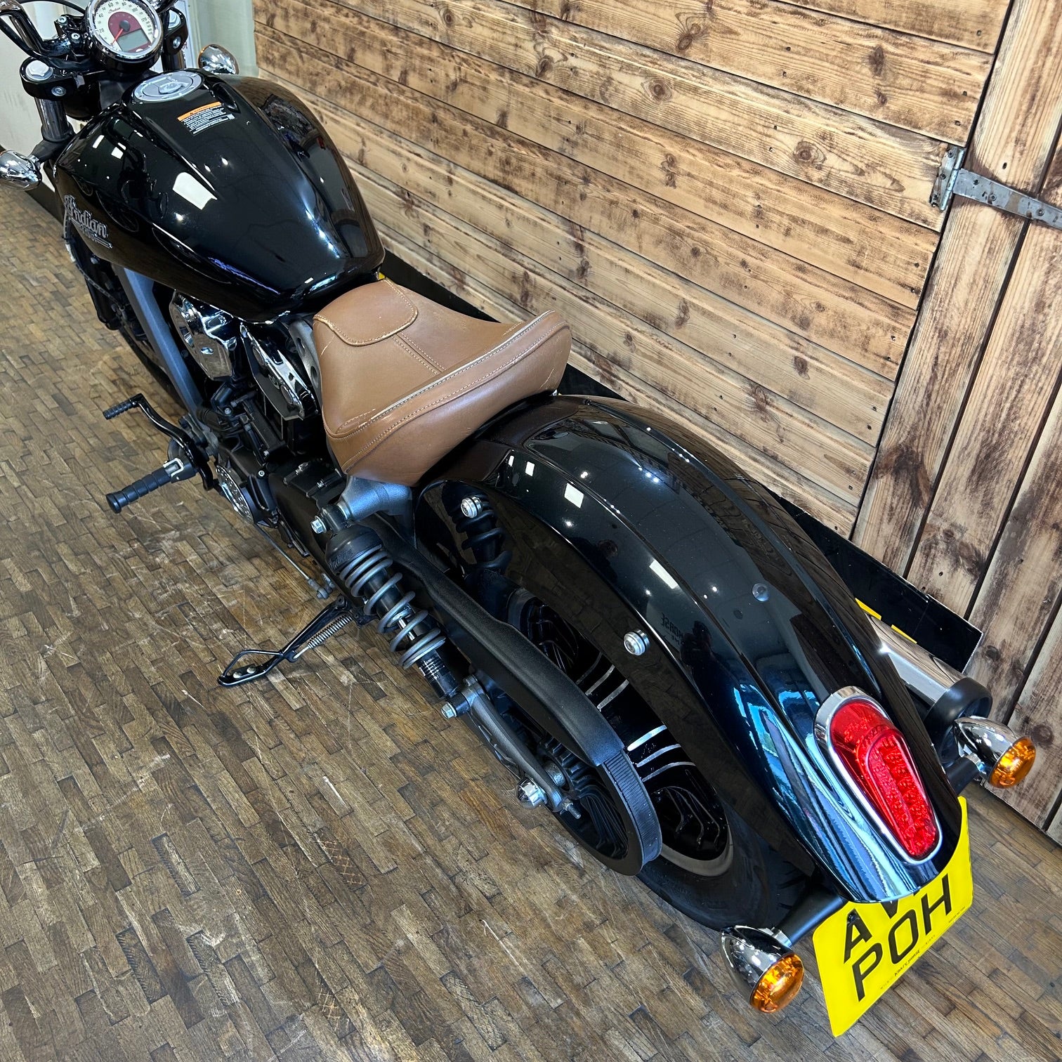Black motorcycle with a brown seat against a wooden wall.