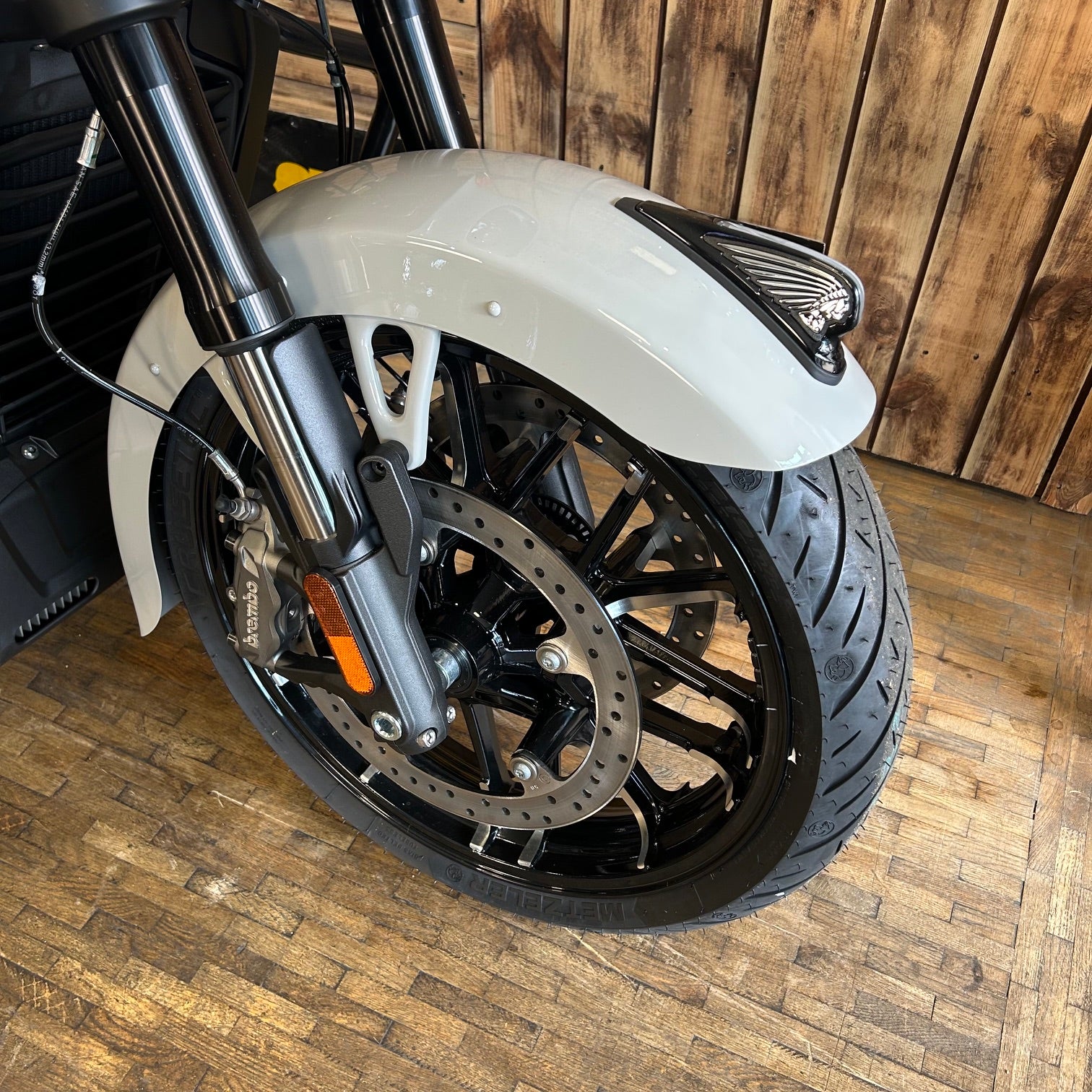 Close-up of a motorcycle wheel with a white fender on a wooden floor.