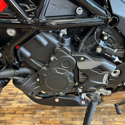 Close-up of a black motorcycle engine and side frame, with visible mechanical components, a wooden floor, and a small metallic badge on the side.