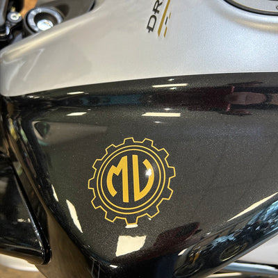 A close-up of a motorcycle tank featuring a gold circular gear emblem with the letters 'MD' in the center, mounted on a glossy black section of the fuel tank. The upper portion of the tank is silver with a partial 'DR.