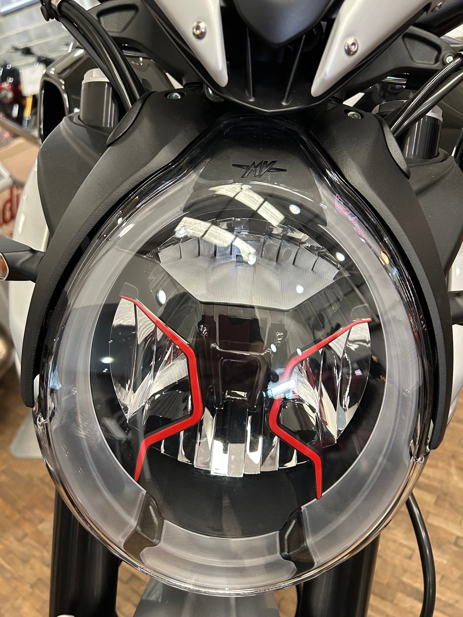 Close-up of a motorcycle headlight with a clear outer lens, black internal structure, and red accent lines, mounted on a black and silver motorcycle.