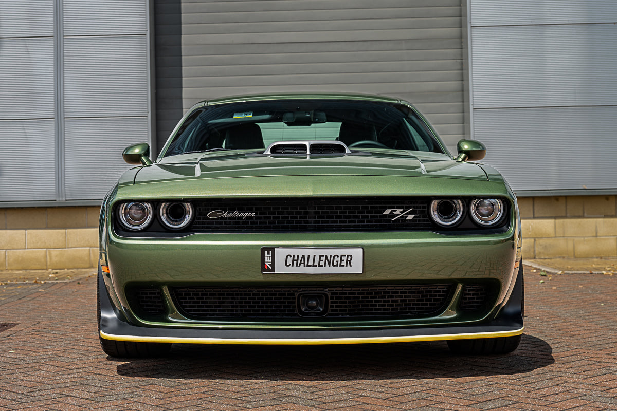 Green Dodge Challenger car parked on a brick driveway with a metallic building in the background.