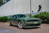 Green Dodge muscle car parked on a brick driveway with a building and trees in the background