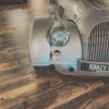 Close-up of a vintage car's front grille and headlight on a wooden floor.