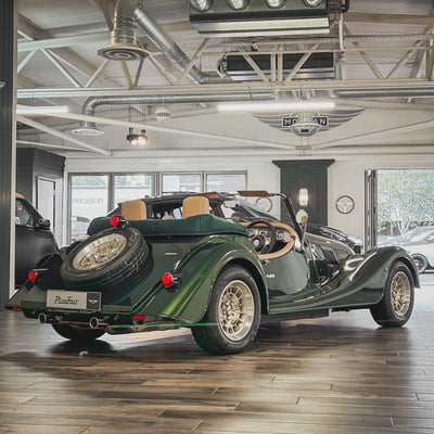 A dark green classic convertible sports car with a spare tire mounted on the rear door, parked inside a spacious showroom with wood floors and industrial ceiling details.