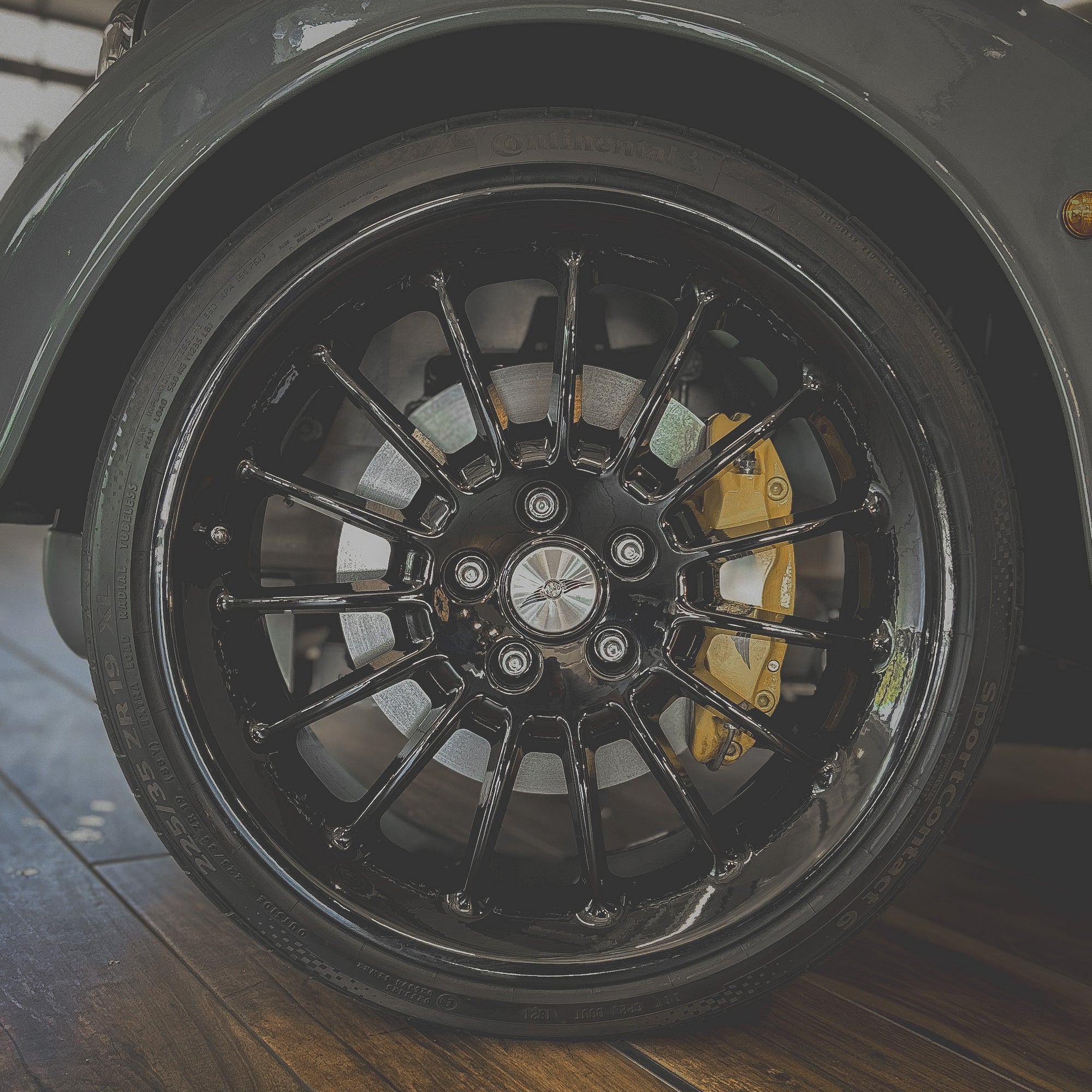 Close-up of a black car wheel with yellow brake caliper on a wooden floor.