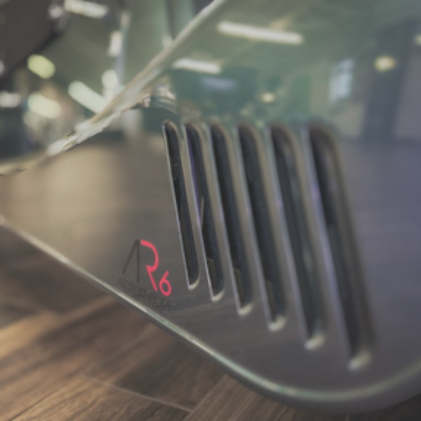 Close-up of a dark gray metallic device with vertical vent-like slits and a small red 'R6' logo, photographed on a wooden floor with a blurred indoor background.