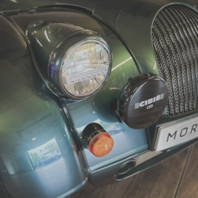 Close-up of a car front end showing a round headlight, a black CIBI LED light mounted on the bumper, a small amber indicator, and part of a dark grille with a license plate reading MOR.