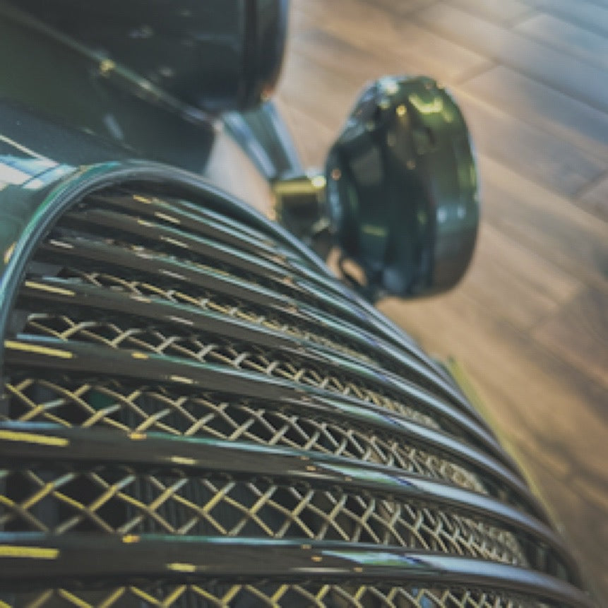 Close-up of a vintage car's chrome grille and round headlight, photographed on a wooden floor.