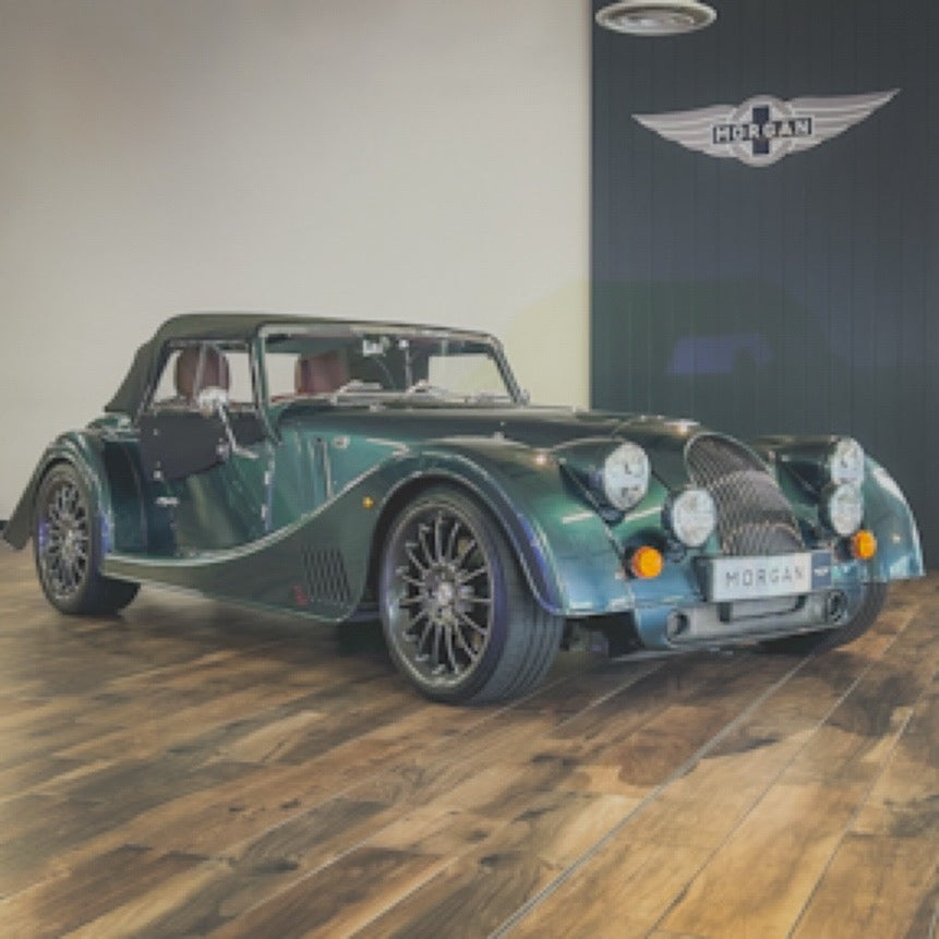 A dark green Morgan Super Sport convertible roadster car displayed indoors on a wooden floor, with a Morgan wing logo visible on the wall behind it.