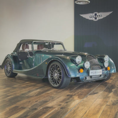 A dark green Morgan Super Sport convertible roadster car displayed indoors on a wooden floor, with a Morgan wing logo visible on the wall behind it.