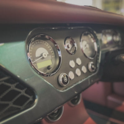 Close-up of a vintage car dashboard featuring round analog gauges, chrome bezels, and a cluster of circular control buttons, with a blurred interior background.