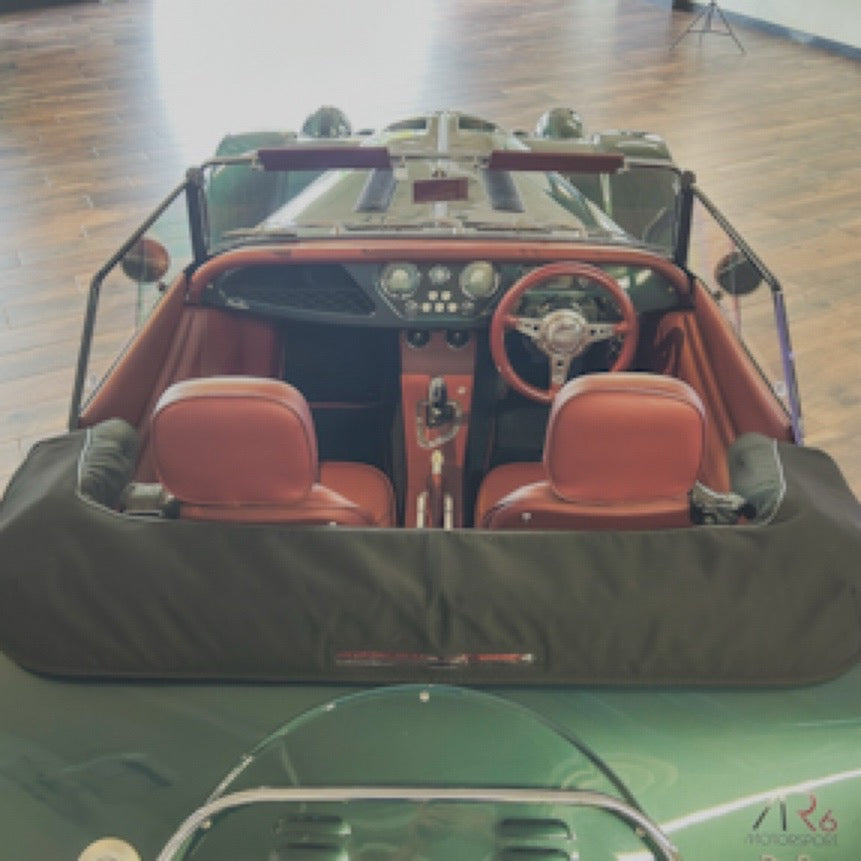 A vintage convertible sports car viewed from above, featuring reddish-brown leather seats, a dark dashboard, and a black soft top cover, parked on a polished wood floor.