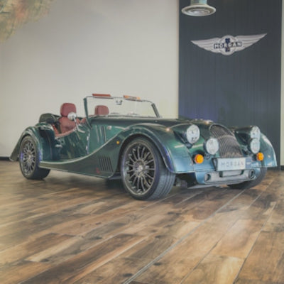 A dark green Morgan convertible sports car with brown leather seats, parked on a wooden floor in front of a wall with a Morgan logo.