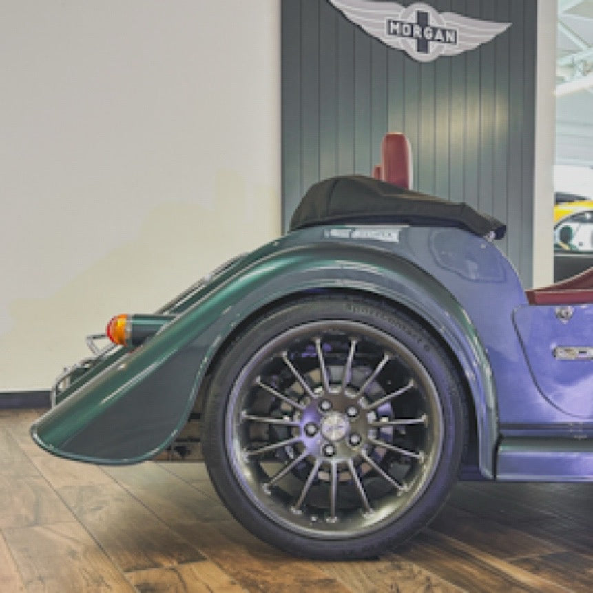 Close-up side view of a vintage-style Morgan car with a dark green fender, black convertible top, and large multi-spoke wheel, parked indoors against a gray paneled wall with a Morgan logo.