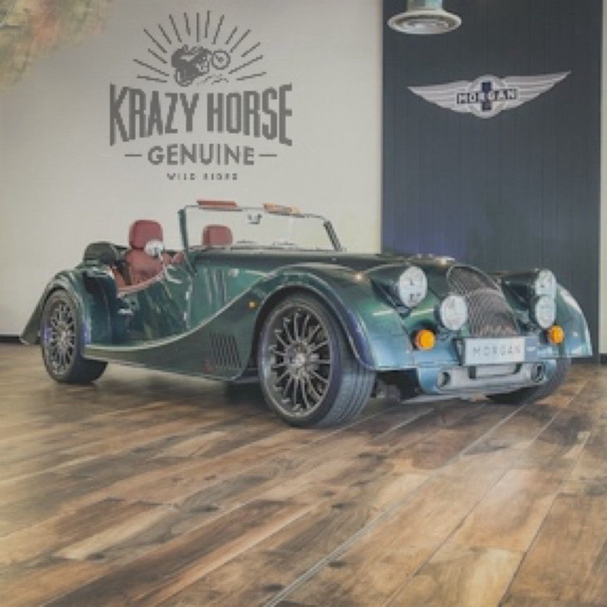 A dark green classic Morgan convertible sports car parked indoors on a wood floor, with wall logos reading 'Krazy Horse Genuine' and 'Morgan' behind it.