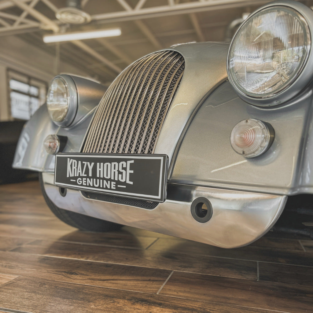 A close-up of a vintage car front end with a black license plate reading 'Krazy Horse Genuine'. The car has a metallic silver-gray body, chrome grille, round headlights, and is photographed indoors on a wood floor.