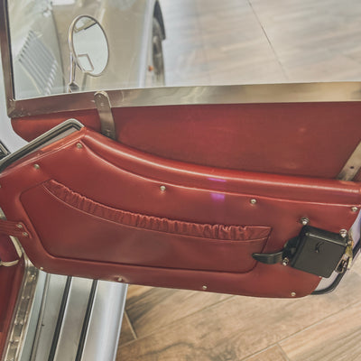 A close-up of a vintage car's interior door panel with reddish-brown vinyl upholstery, a fabric pocket, and silver metal trim, shown with part of the car door frame and wood floor visible.