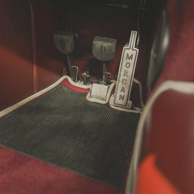 A vintage-style metal clutch pedal with the word 'MORGAN' printed vertically, installed in a car footwell with red interior trim and a black ribbed floor mat.