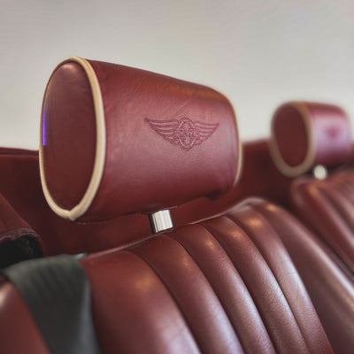 Close-up of a car seat headrest in deep burgundy leather with a debossed winged emblem, showing the seat cushion and another headrest in the background.