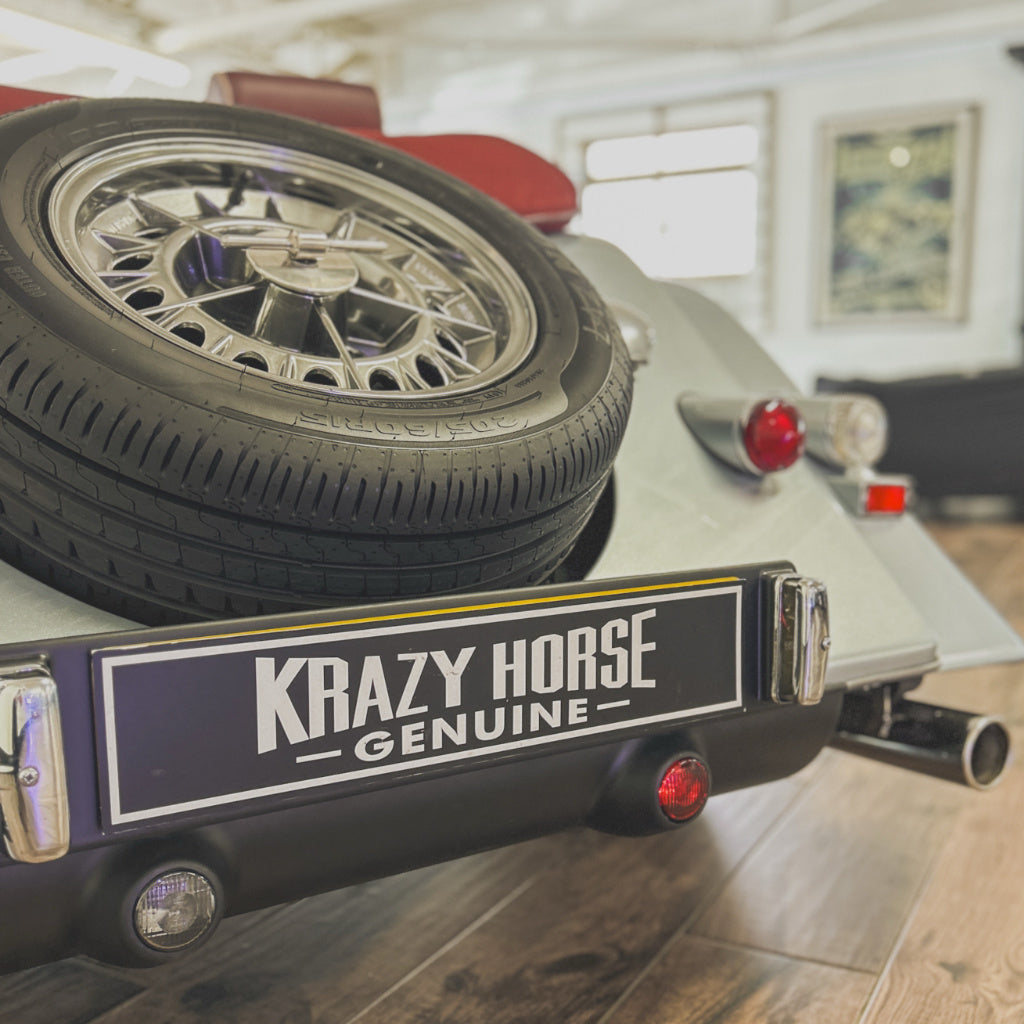 A close-up of a vintage-style vehicle rear end with a spare tire mounted on the back, a black bumper, and a sign reading 'KRAZY HORSE GENUINE' in white lettering. The photo is taken indoors on a wood floor with a blurred interior background.
