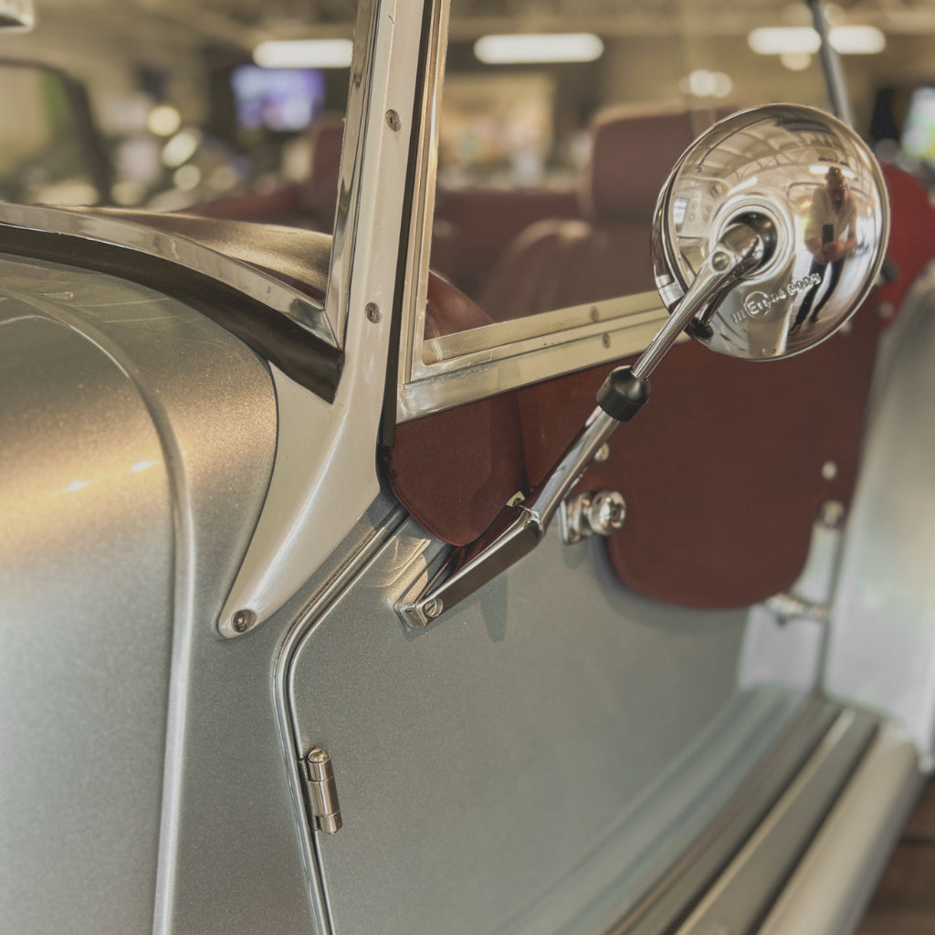 Close-up of a vintage car door with a chrome side mirror, silver body trim, and reddish-brown interior upholstery in a showroom setting.