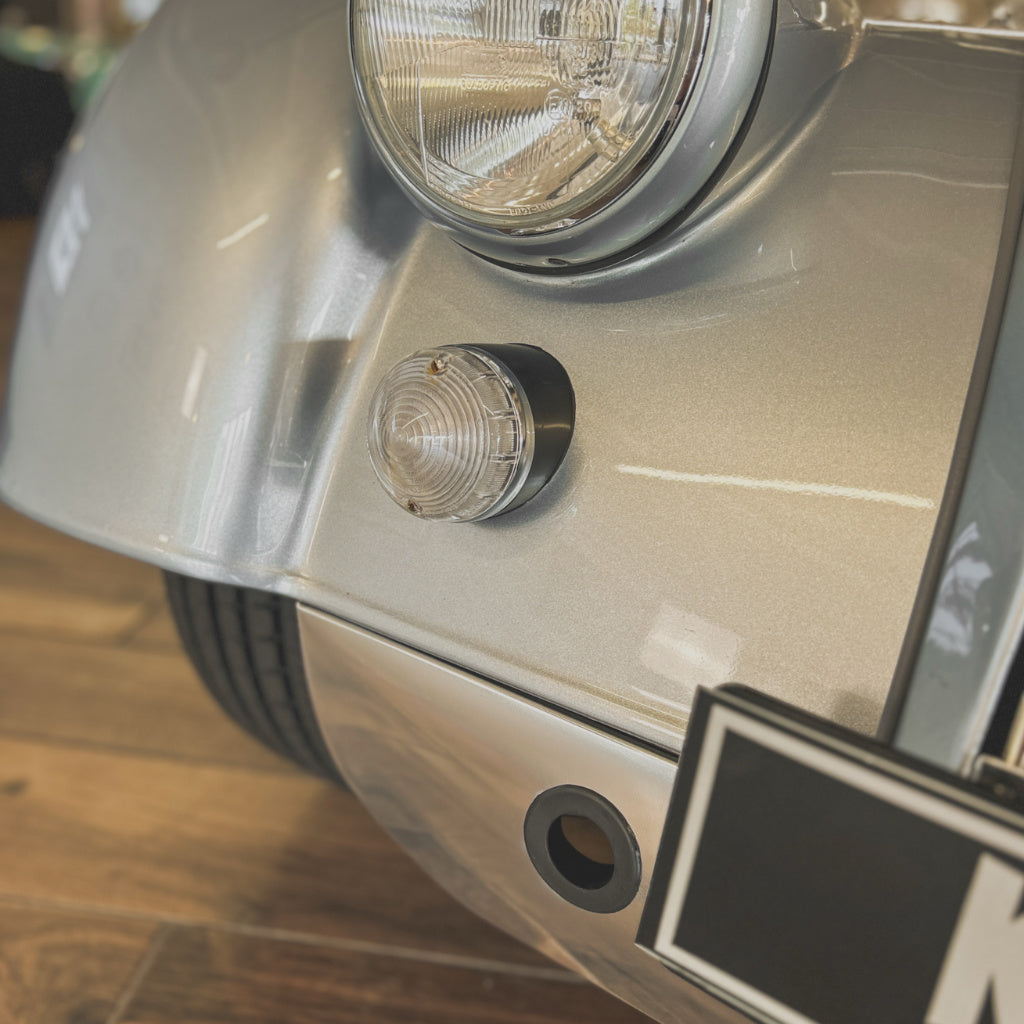 Close-up of a silver car body panel showing a round clear marker light, headlight, tire, and part of a license plate, photographed indoors on a wood floor.